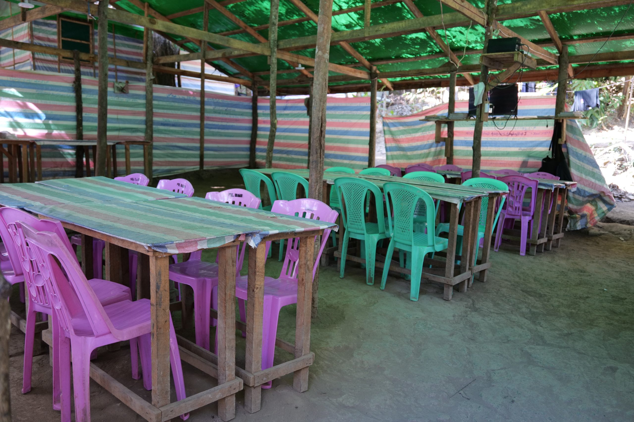Colorful plastic chairs and wooden tables are arranged in a rustic outdoor dining area with a green canopy and striped walls.