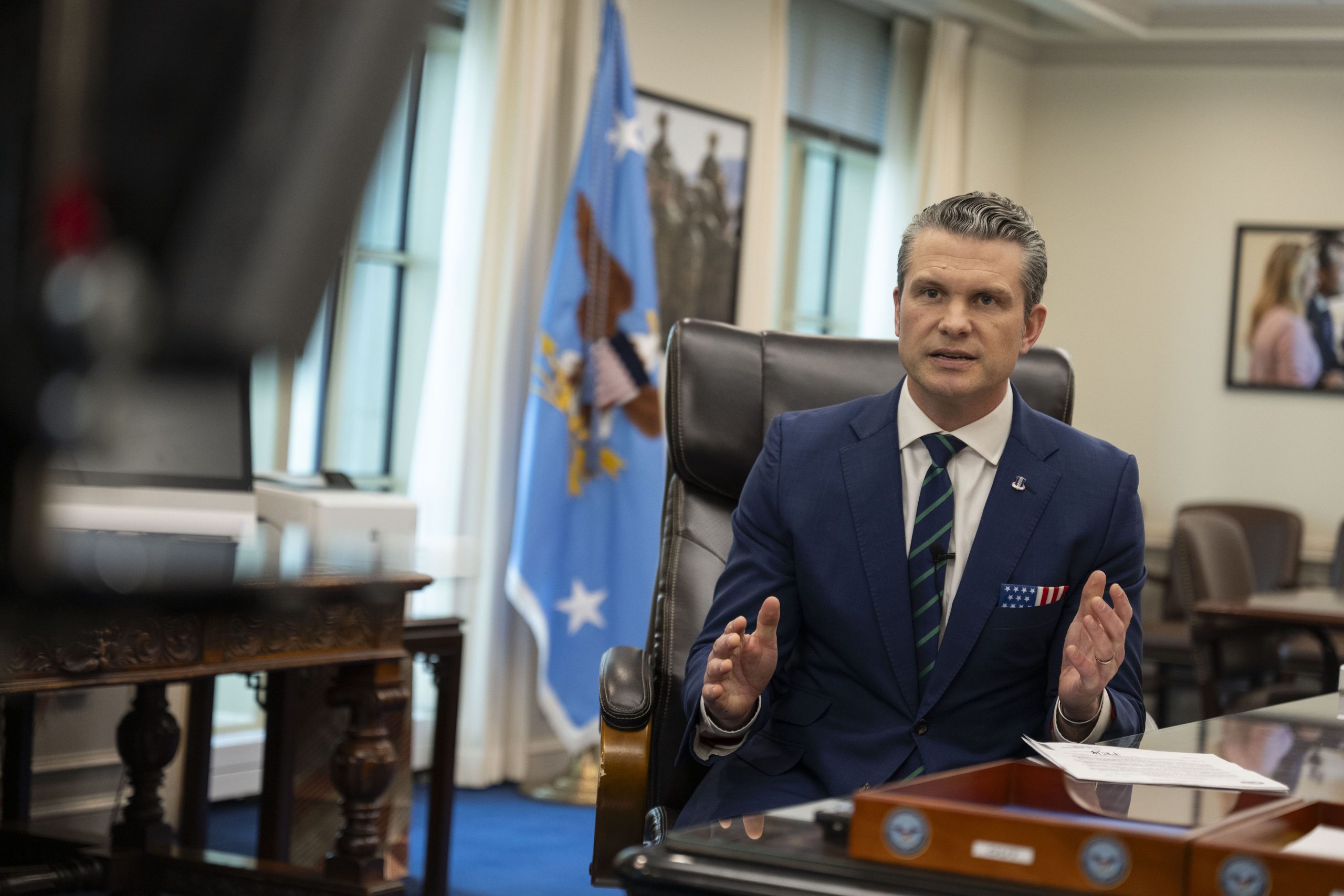 Official in a blue suit gestures while speaking in an office setting, with a U.S. flag and military-themed decor in the background.