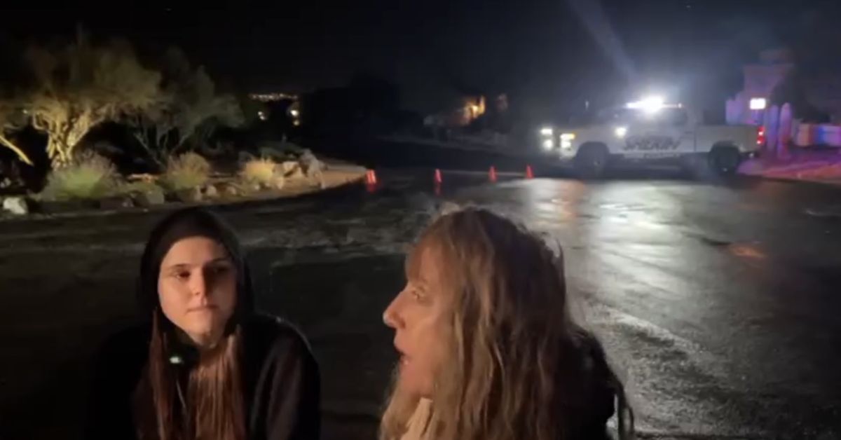 Two individuals converse at night near a police vehicle, with illuminated cones marking off the area in a dimly lit setting.
