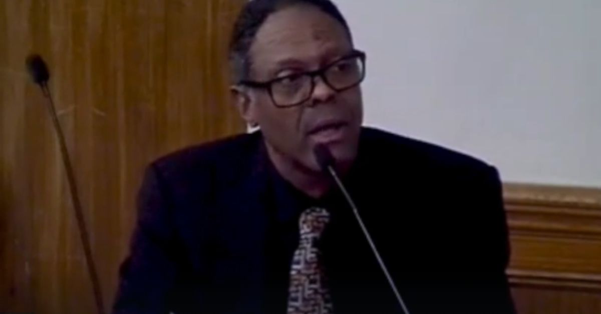 Man speaking at a podium in a formal setting, wearing glasses and a black suit with a patterned tie, engaged in a serious discussion.
