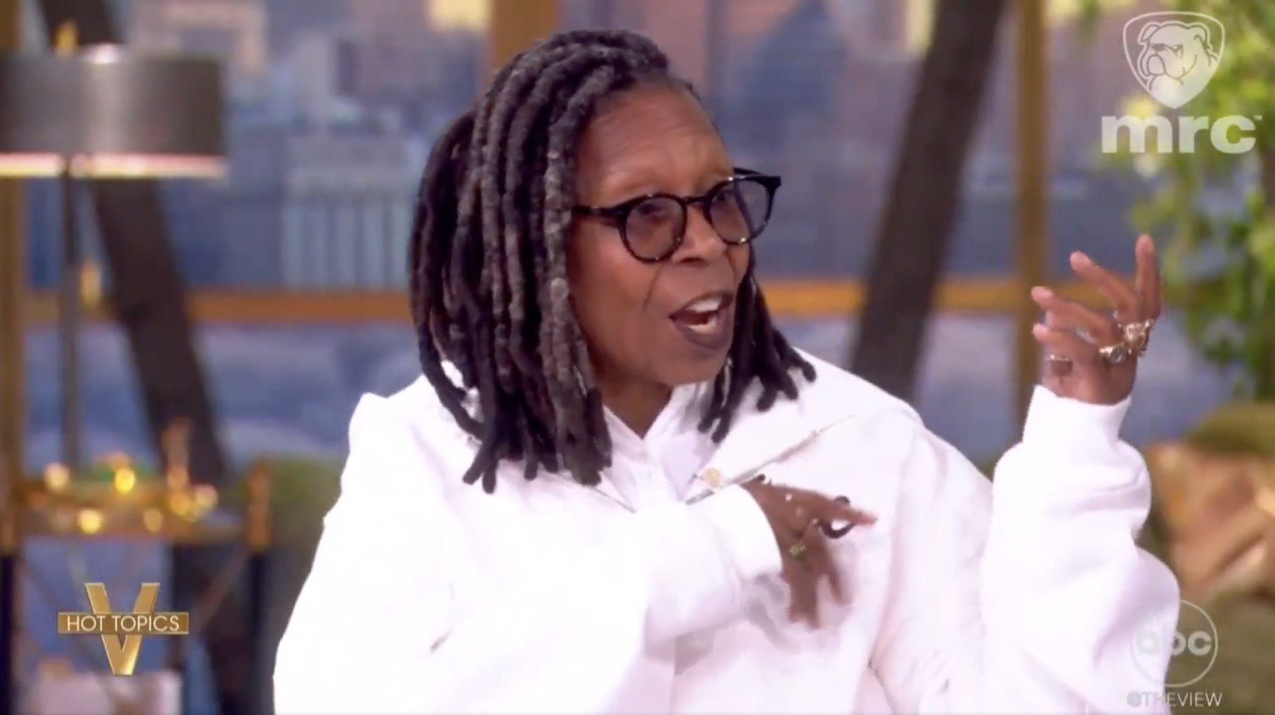 Whoopi Goldberg discussing current events on a talk show set, wearing a white jacket and glasses, with a city skyline in the background.