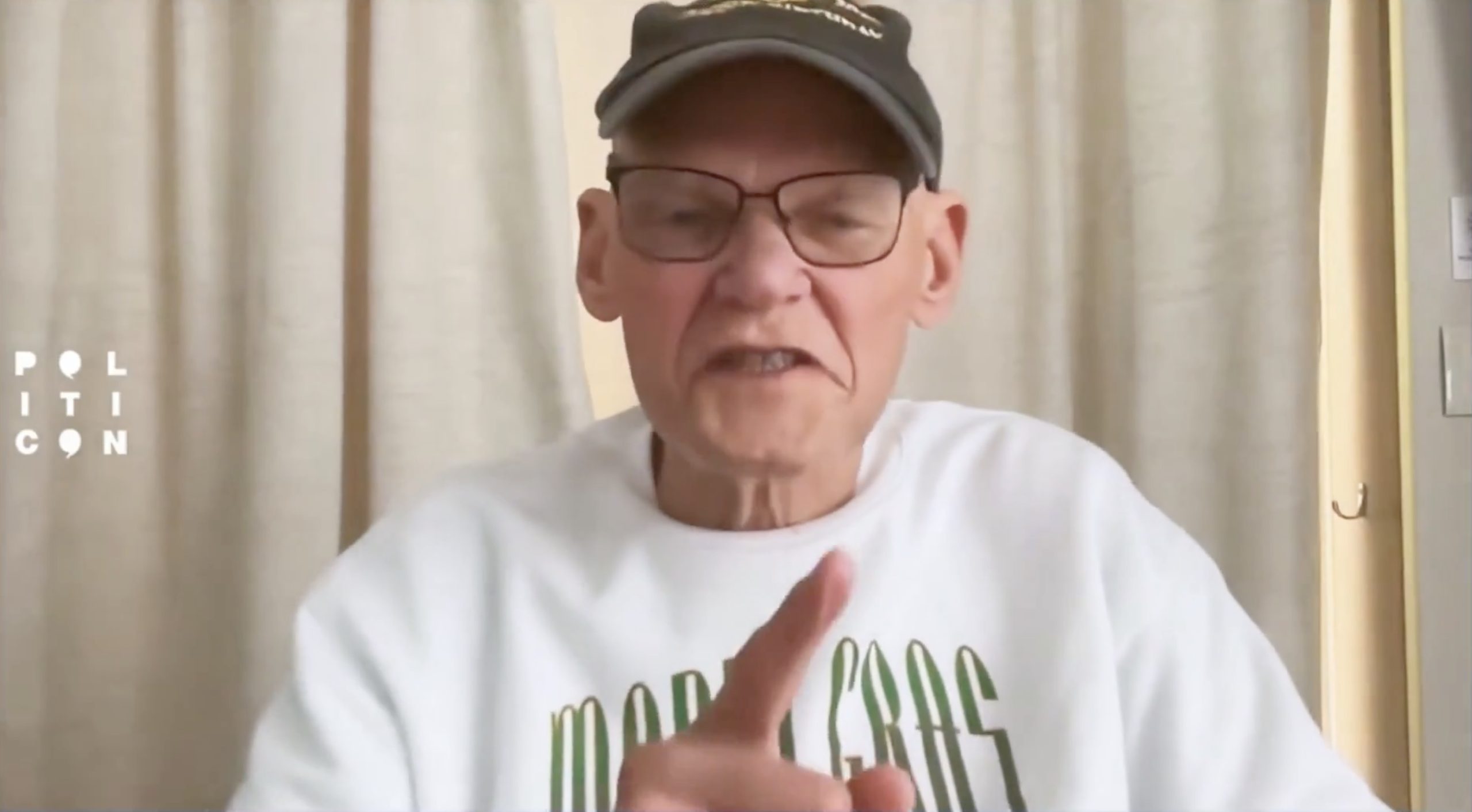 James Carville speaking passionately during a Politicon event, wearing a cap and a white shirt, gesturing with his hand.