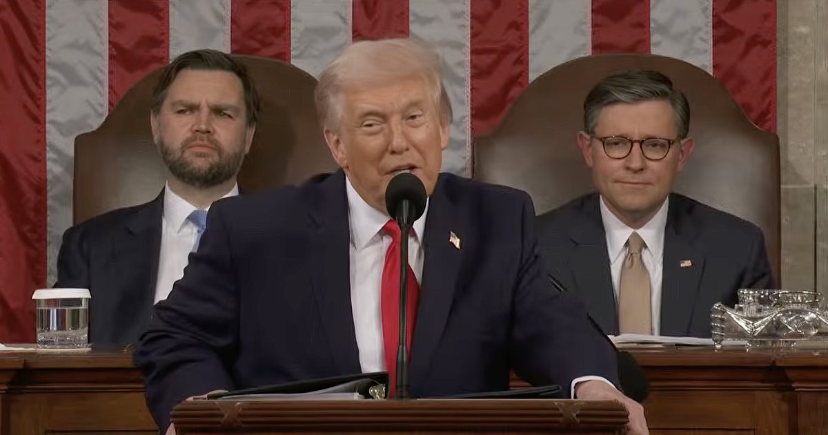 Donald Trump speaking at a podium during a formal address, flanked by two seated officials, with an American flag backdrop.