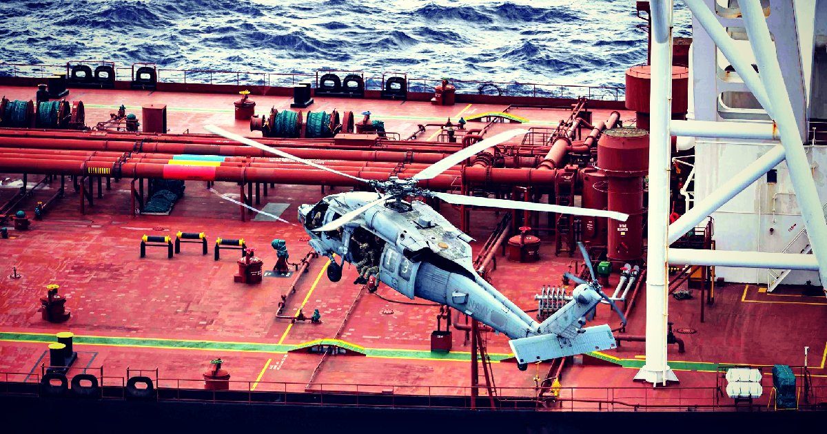 A helicopter hovers above a large cargo ship, with visible pipelines and equipment on the deck, against a backdrop of ocean waves.