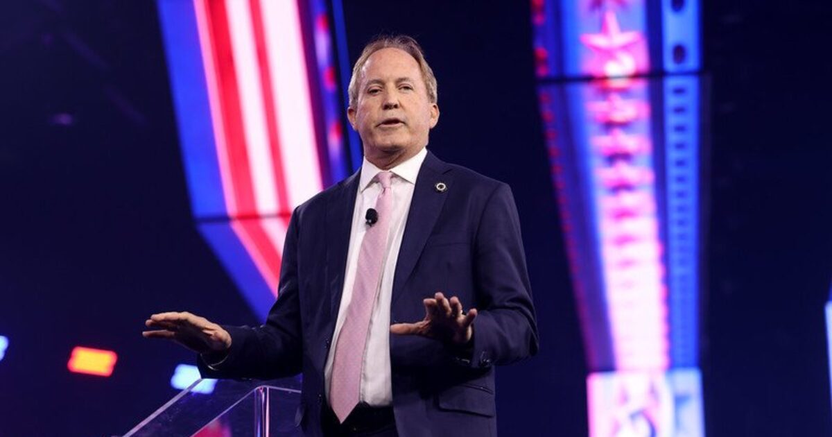 Speaker addressing an audience at a conference, wearing a suit and tie, with colorful stage lighting in the background.