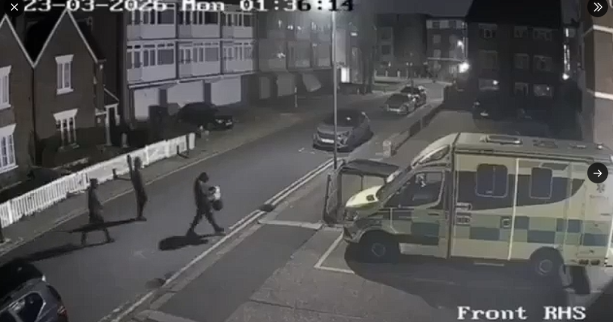 Three individuals walk along a dimly lit street near an ambulance parked on the side, captured by a security camera at night.