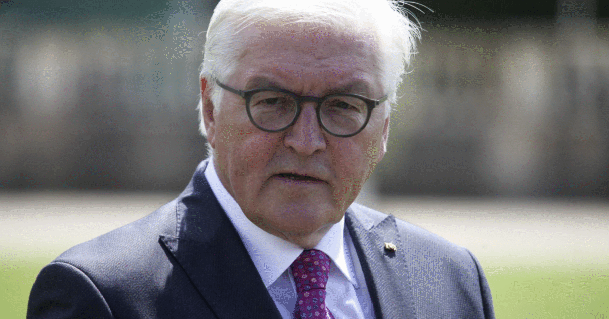 German politician with white hair and glasses wearing a navy suit and pink tie, standing outdoors in a public setting.