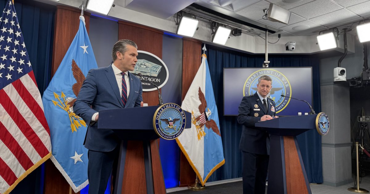 Press conference at the Pentagon featuring two officials, one in a suit and the other in military uniform, with American and Air Force flags in the background.