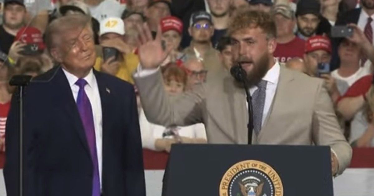 Donald Trump and Jake Paul speaking at a rally, with an engaged audience wearing hats and holding signs in the background.