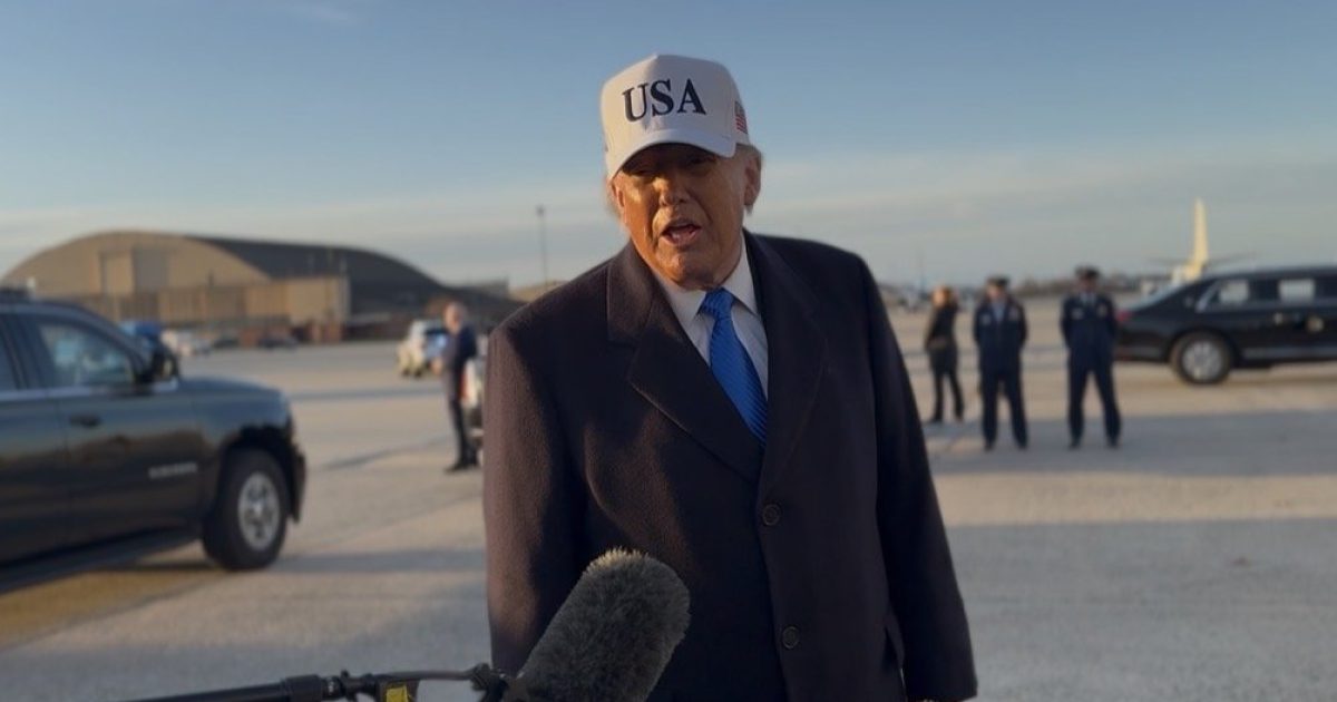 Former President Donald Trump speaks to the media while wearing a USA cap at an airport, with vehicles and military personnel visible in the background.