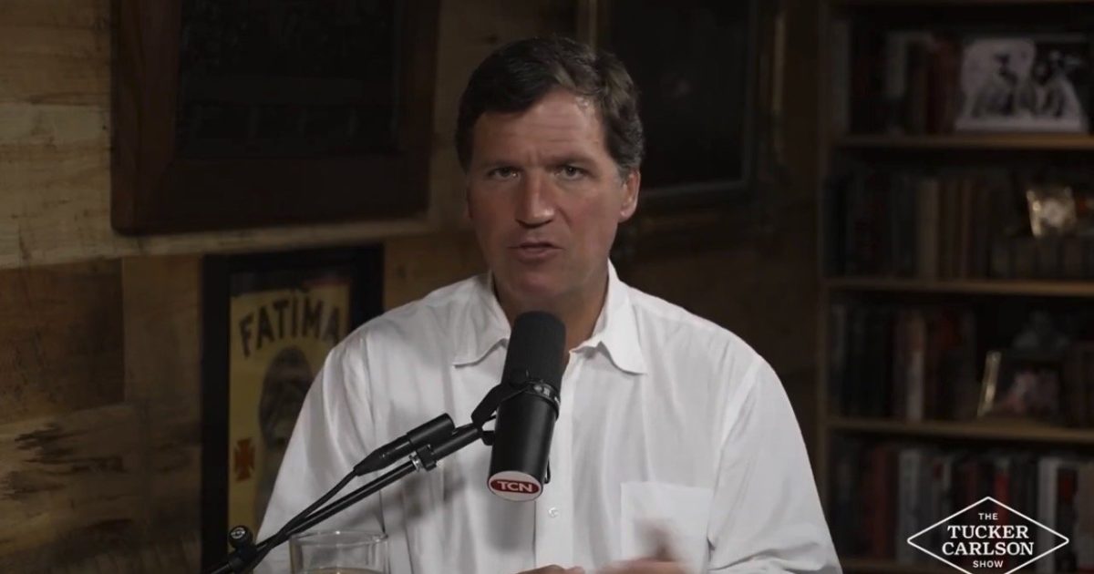 Tucker Carlson discussing current events on his show, seated at a desk with a microphone and a glass, in a cozy, wood-paneled setting.
