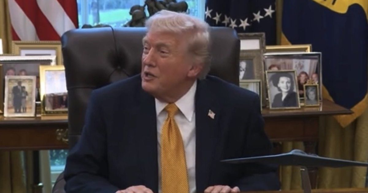 Donald Trump seated in the Oval Office, smiling during a discussion, with a backdrop of American flags and personal photographs on the desk.