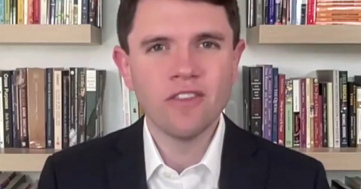Man in a suit smiling in front of a bookshelf filled with various books, showcasing a professional and academic environment.