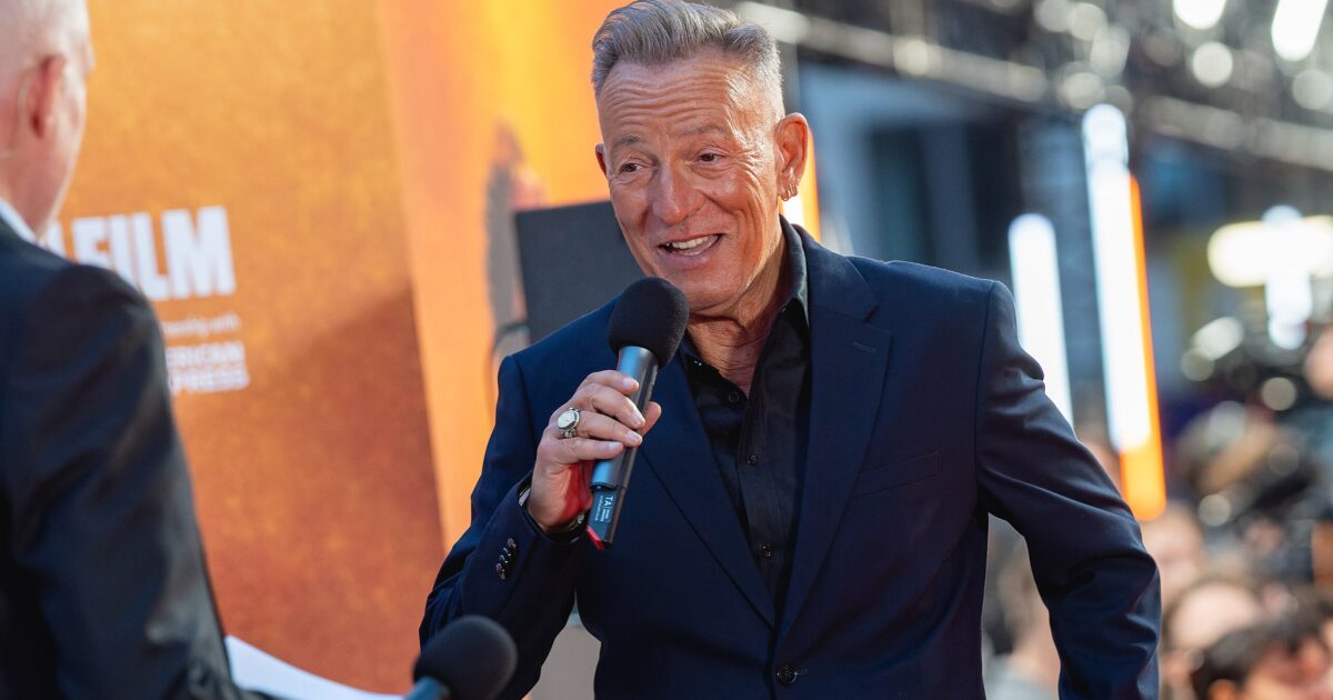 A man in a black suit speaks into a microphone at a film event, with bright lights and an audience in the background.