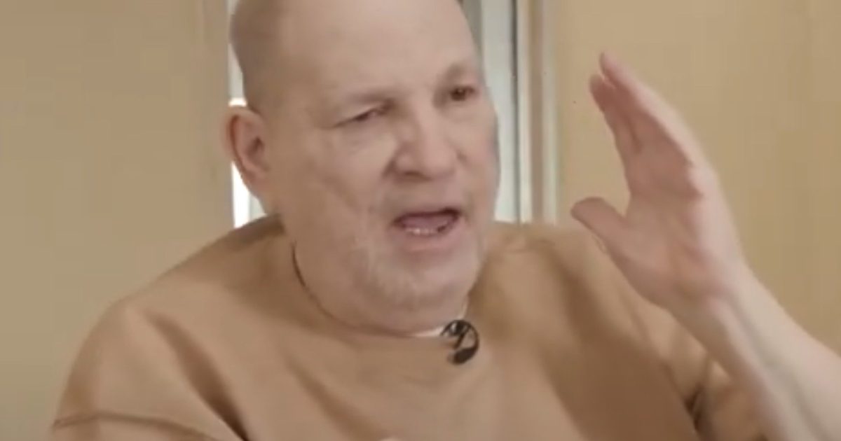 Man in a beige sweater gestures while speaking, expressing emotions during an interview in a well-lit indoor setting.