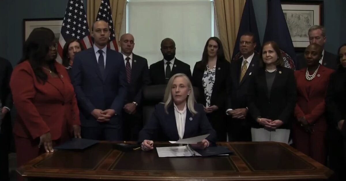 A woman in formal attire sits at a desk with paperwork, surrounded by officials and flags, during a governmental signing event.