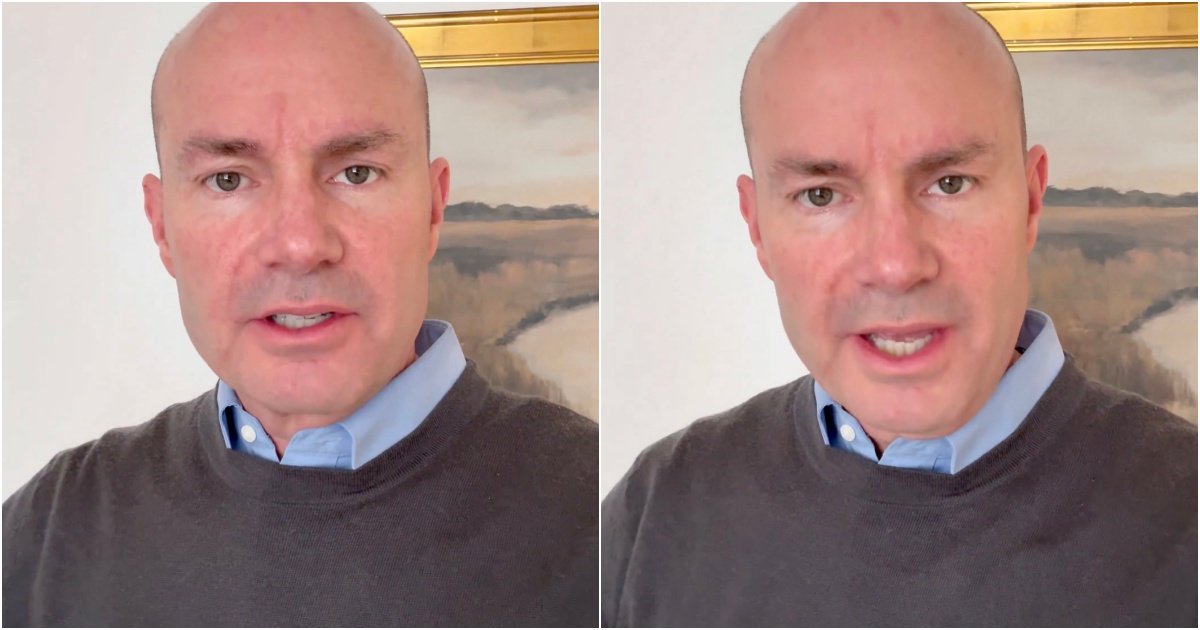 Man speaking directly to the camera while wearing a gray sweater and blue shirt, with a landscape painting in the background.