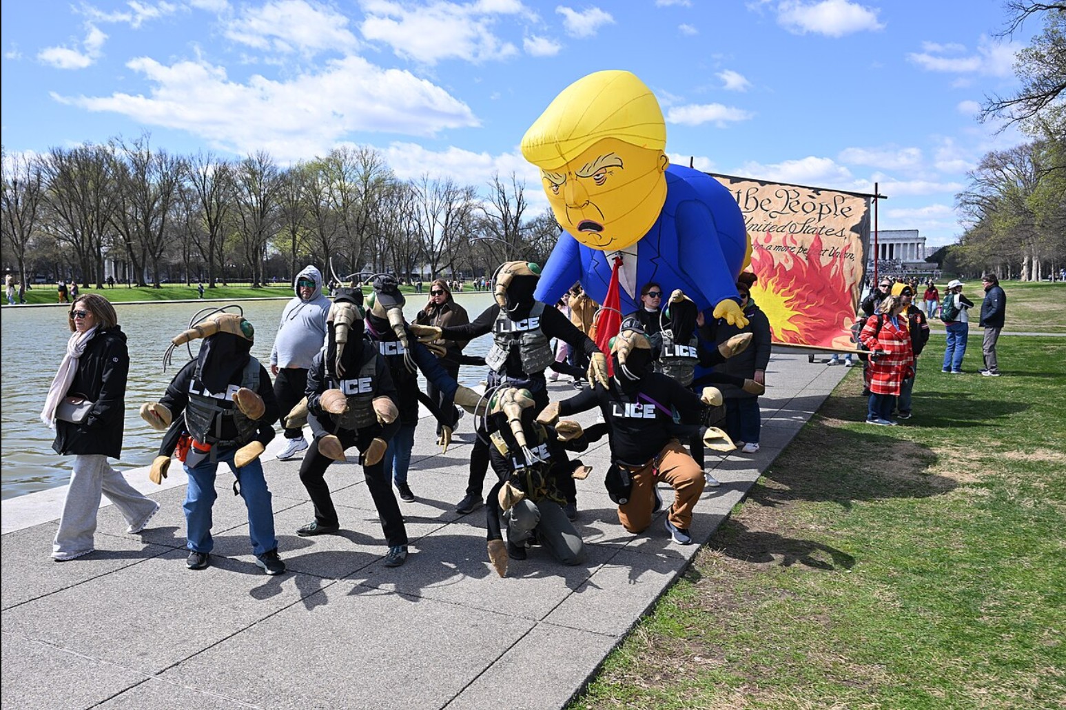 Protesters in costumes representing ICE pose in front of a large inflatable Trump figure during a demonstration at a public park.