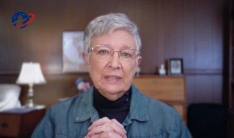 Elderly woman with short gray hair and glasses speaking in a cozy room, conveying a message of importance and sincerity.