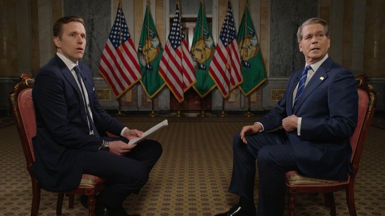 Interview setting featuring two men seated, one holding notes, with U.S. flags and green banners in the background.