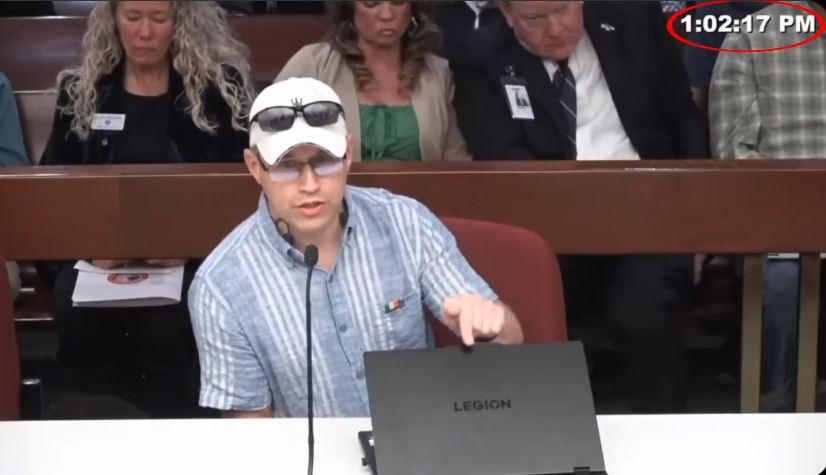 Man in a blue plaid shirt speaks at a public meeting, presenting information on a laptop while an audience listens attentively in the background.