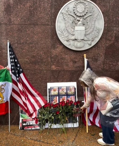 A woman places roses at a memorial site featuring American and Mexican flags, with a backdrop of a seal and protest signs.