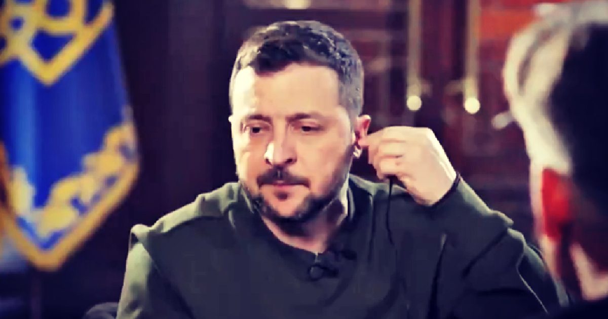 Ukrainian leader engaged in conversation while adjusting an earpiece, with a national flag in the background.