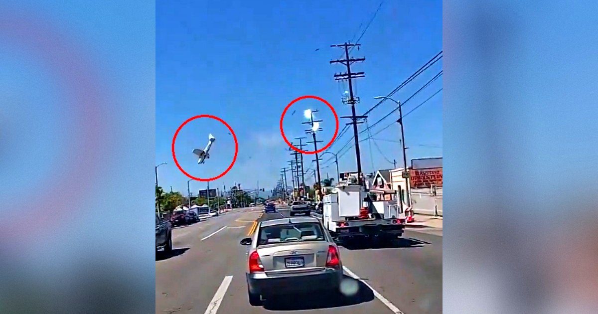 A small airplane is seen flying upside down above a busy street, with vehicles and power lines visible below in clear blue skies.