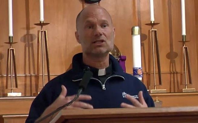 Clergy member delivering a sermon in a church setting, gesturing while speaking to the congregation.