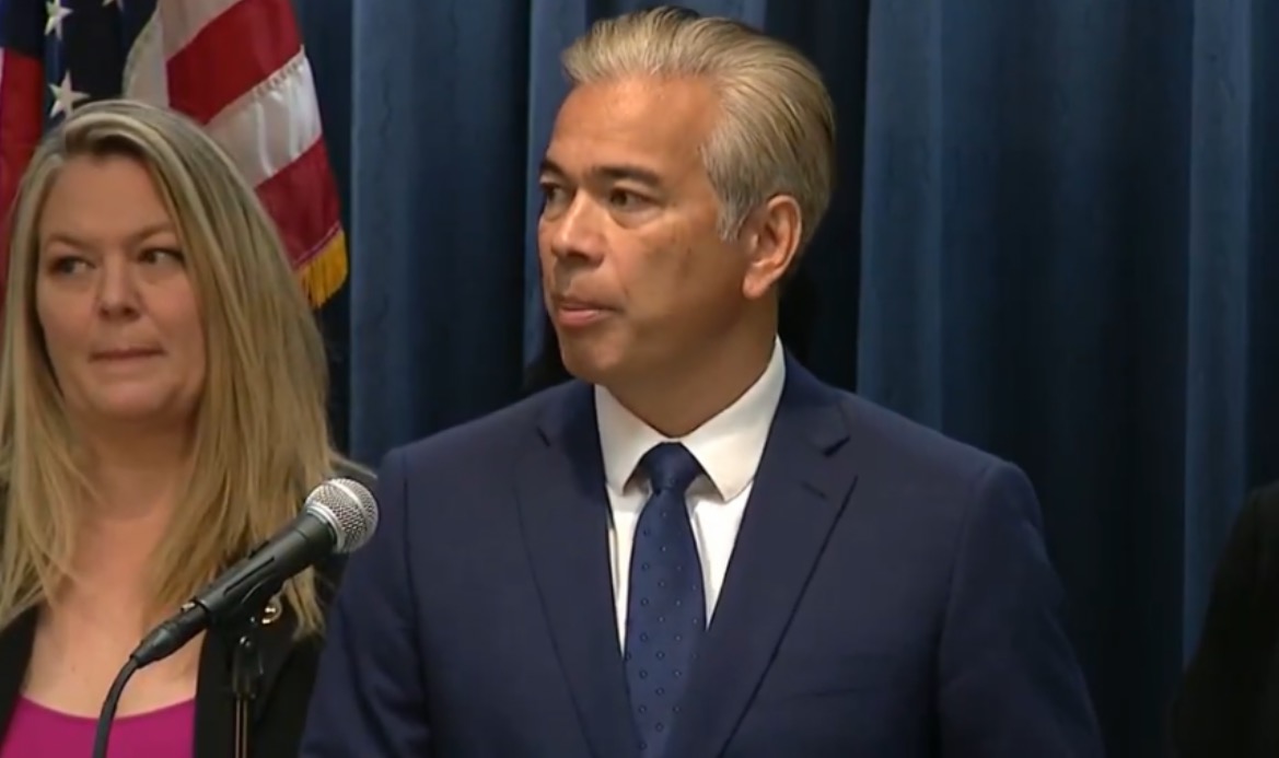 Press conference featuring a speaker in a suit and tie, with a microphone and American flag in the background, discussing important issues.
