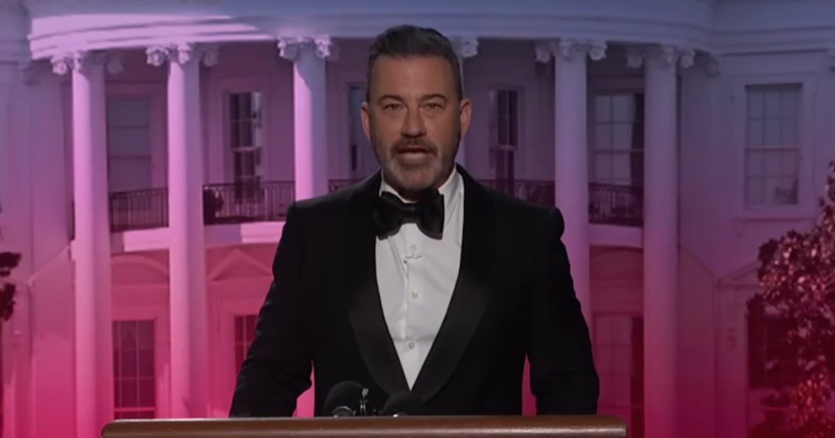 Jimmy Kimmel speaking at the White House Correspondents' Dinner, wearing a tuxedo, with the White House backdrop illuminated in pink.