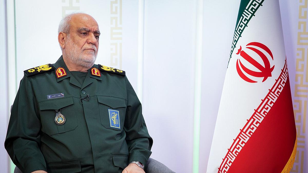 Iranian military official in uniform sitting beside the Iranian flag, conveying a serious expression during an interview or press conference.
