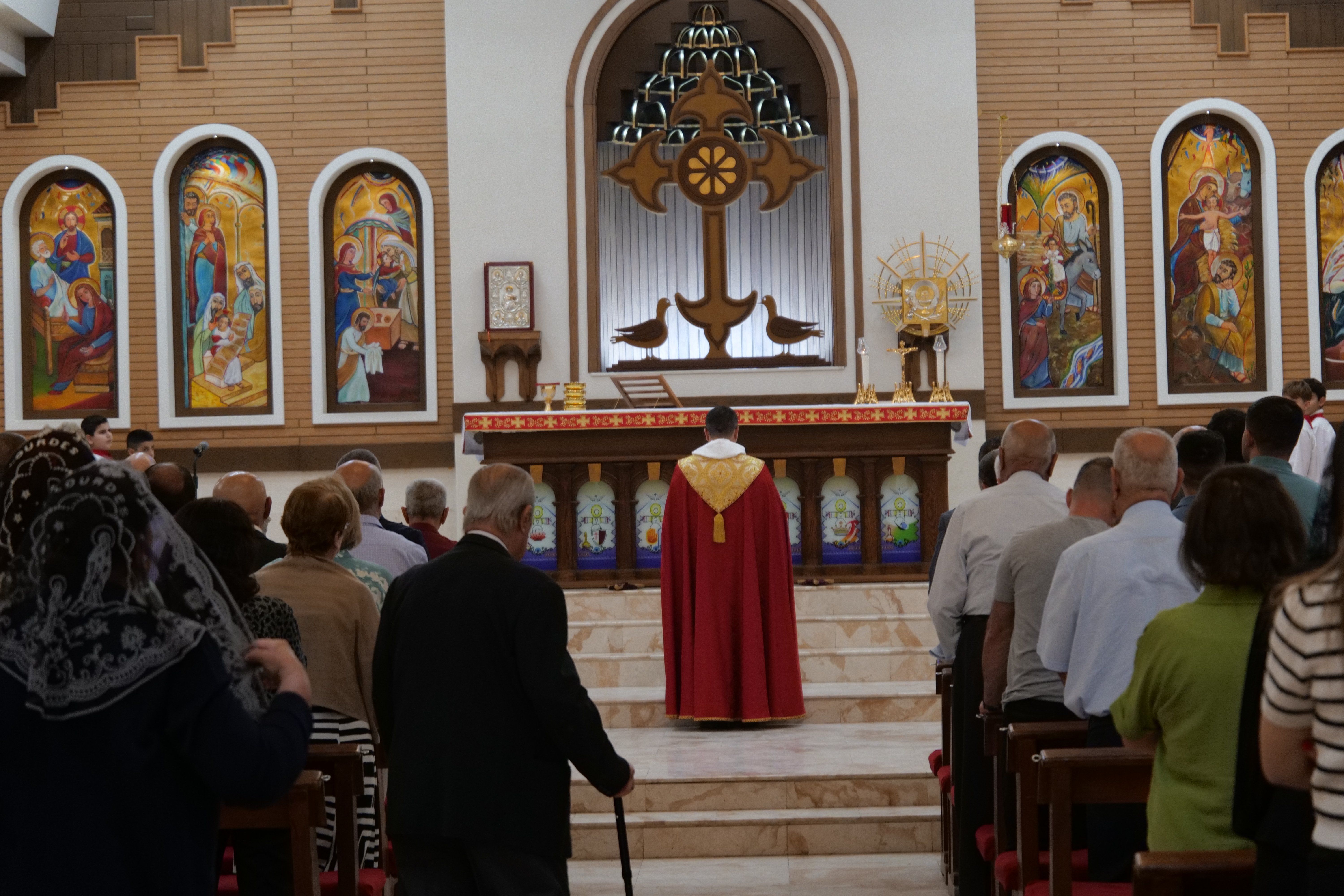 Congregation attending a religious service in a church, featuring an altar with ornate decorations and vibrant stained glass artwork.