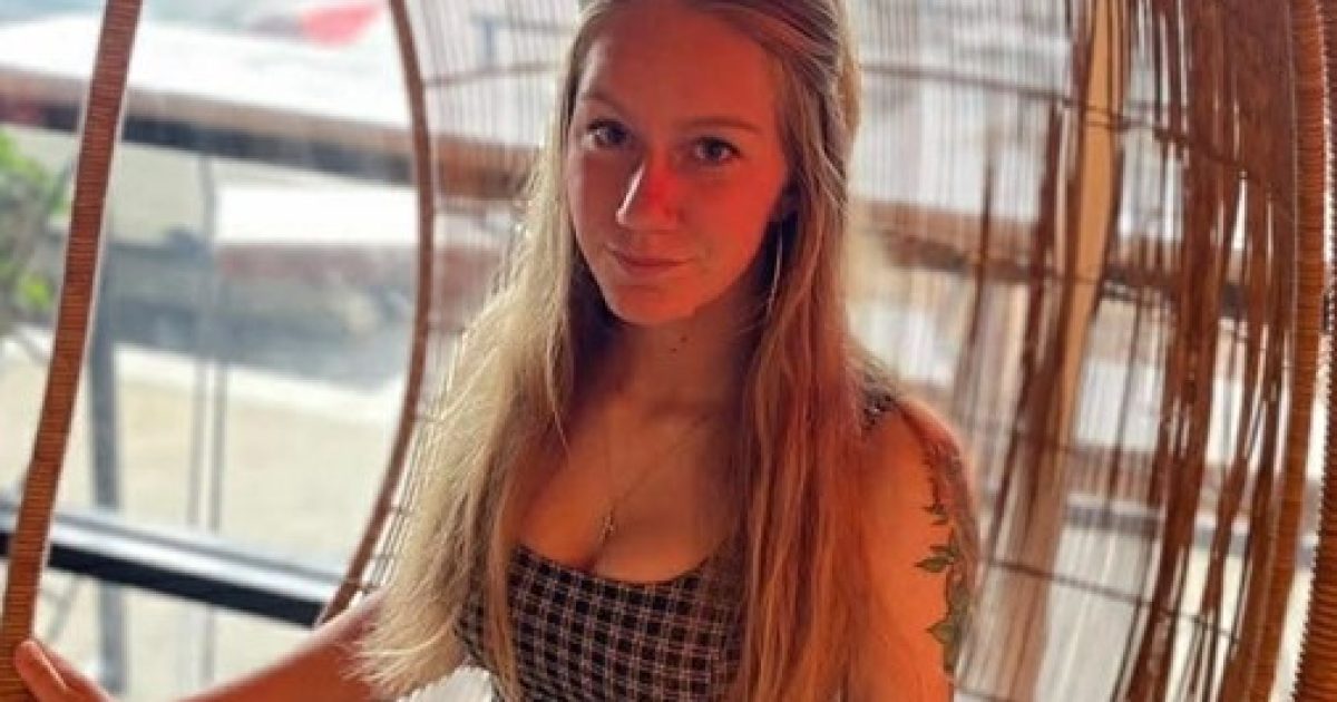 Young woman with long hair sitting in a woven chair, wearing a checkered top, in a cozy indoor setting.
