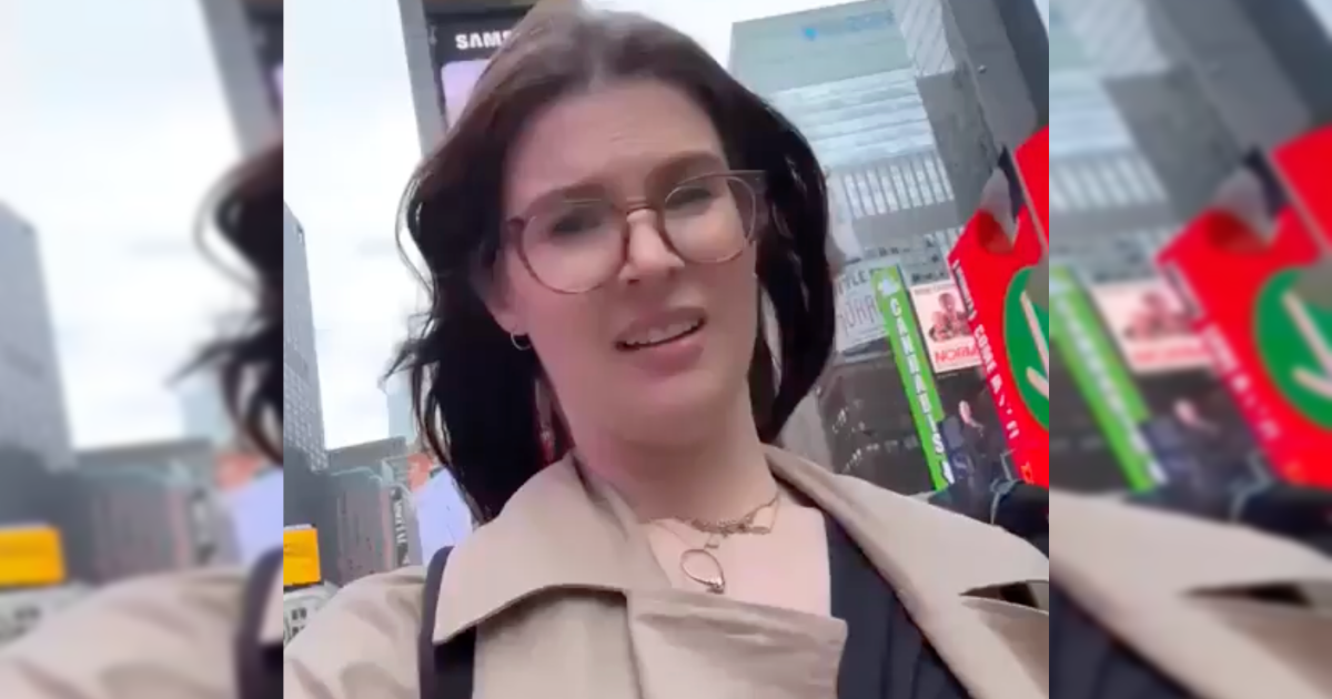 Woman with glasses walking in Times Square, surrounded by vibrant advertisements and city skyscrapers on a cloudy day.
