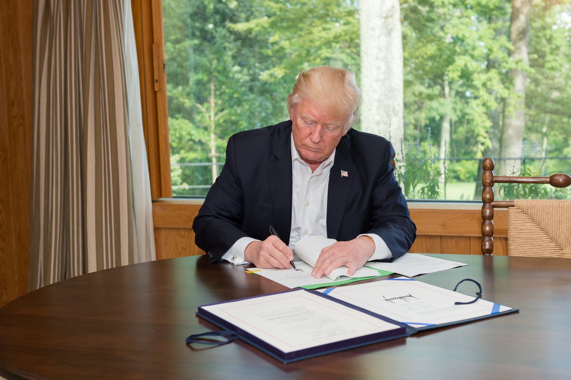 Donald Trump signing documents at a table with a view of greenery in the background.