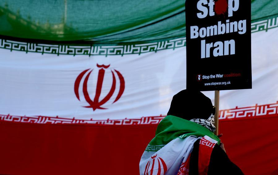 Protester holding a sign reading "Stop Bombing Iran" in front of the Iranian flag during a peace demonstration.