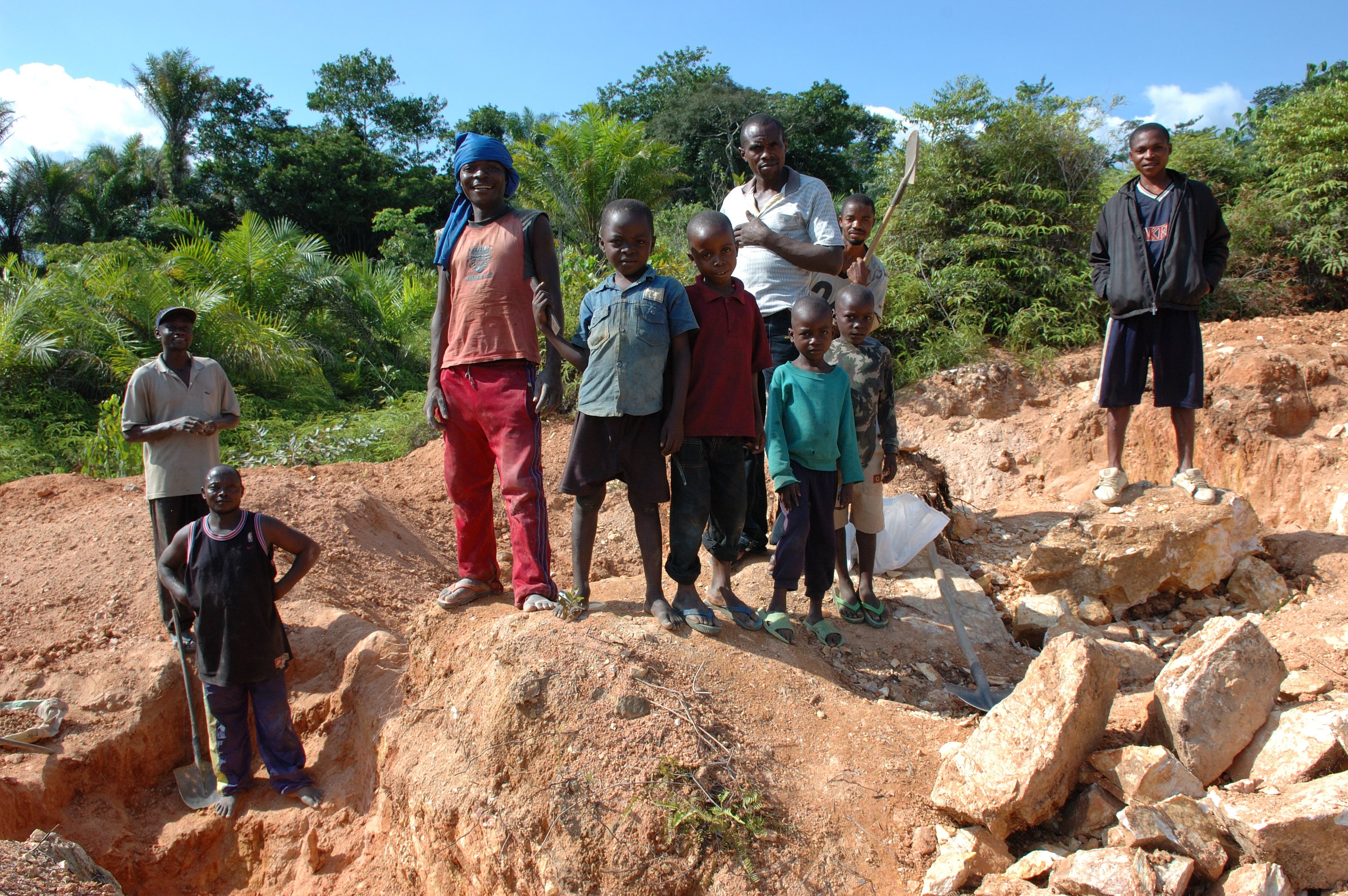 Group of children and adults standing on a mining site surrounded by rocks and vegetation, showcasing community labor and engagement in resource extraction activities.