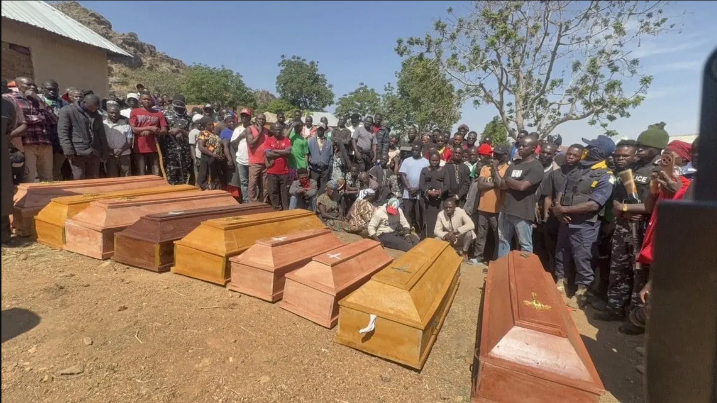 A crowd gathers around wooden coffins during a somber memorial service in a rural area, reflecting a moment of mourning and community support.