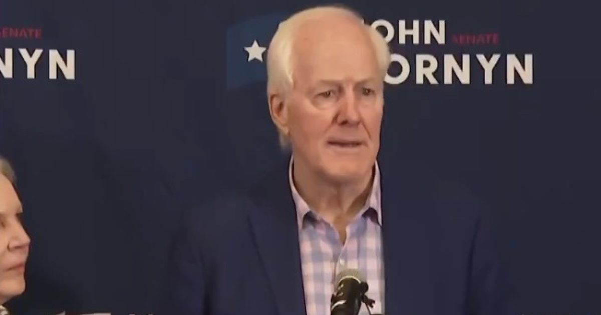 John Cornyn speaking at a press conference, addressing supporters with a blue backdrop featuring his campaign branding.