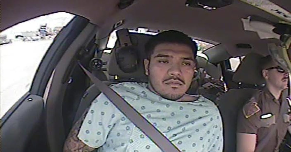 A man in a hospital gown sits in a police car, restrained by a seatbelt, beside a uniformed officer.