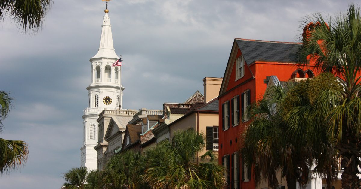 Historic buildings line the waterfront in Charleston, South Carolina, with a prominent white church tower and palm trees under a cloudy sky.