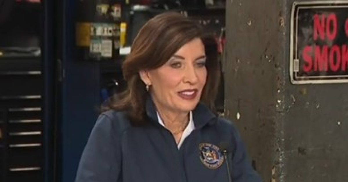 New York Governor Kathy Hochul speaking at a press conference, wearing a dark jacket with the state seal, in a workshop setting.