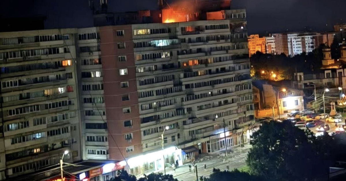 Night view of a residential building with flames visible on the roof, surrounded by city lights and parked cars below.