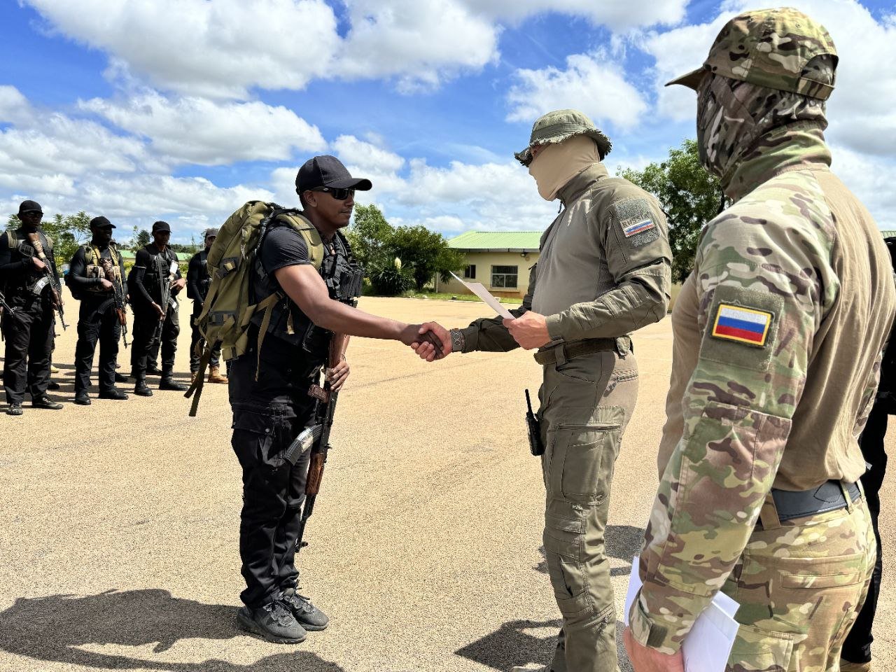 Military personnel engage in a formal handshake during a training exercise, with armed soldiers in the background, highlighting teamwork and collaboration.