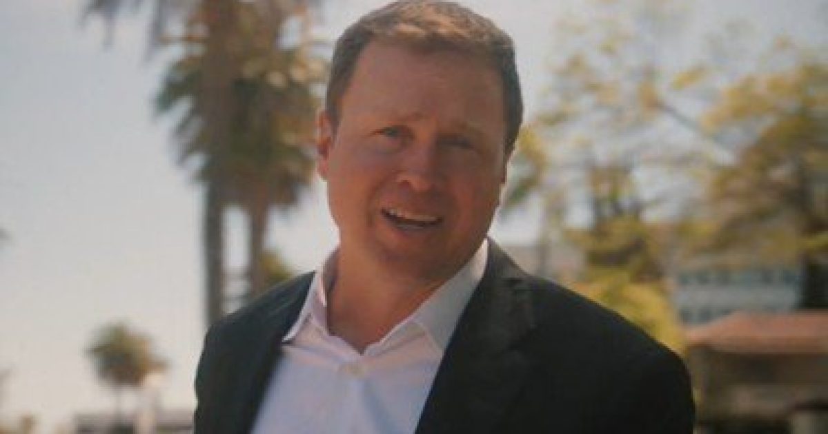 Business professional in a suit standing outdoors with palm trees in the background, conveying a confident demeanor and engaged in conversation.