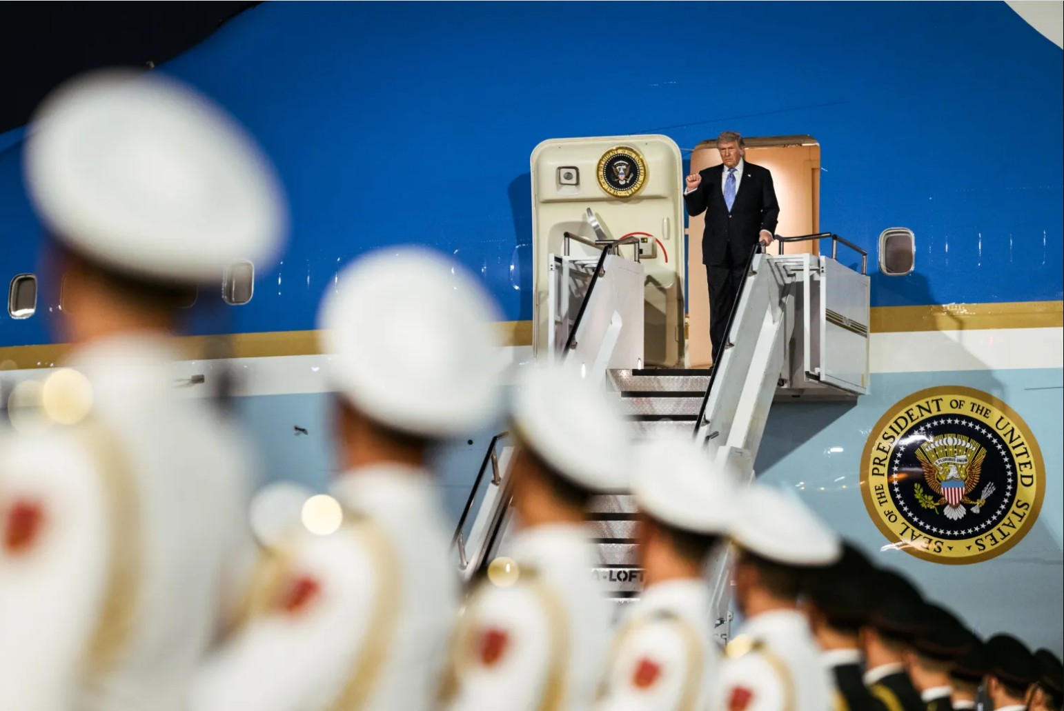 Former President Donald Trump waves from the steps of Air Force One, with a military honor guard in the foreground during a ceremonial arrival.