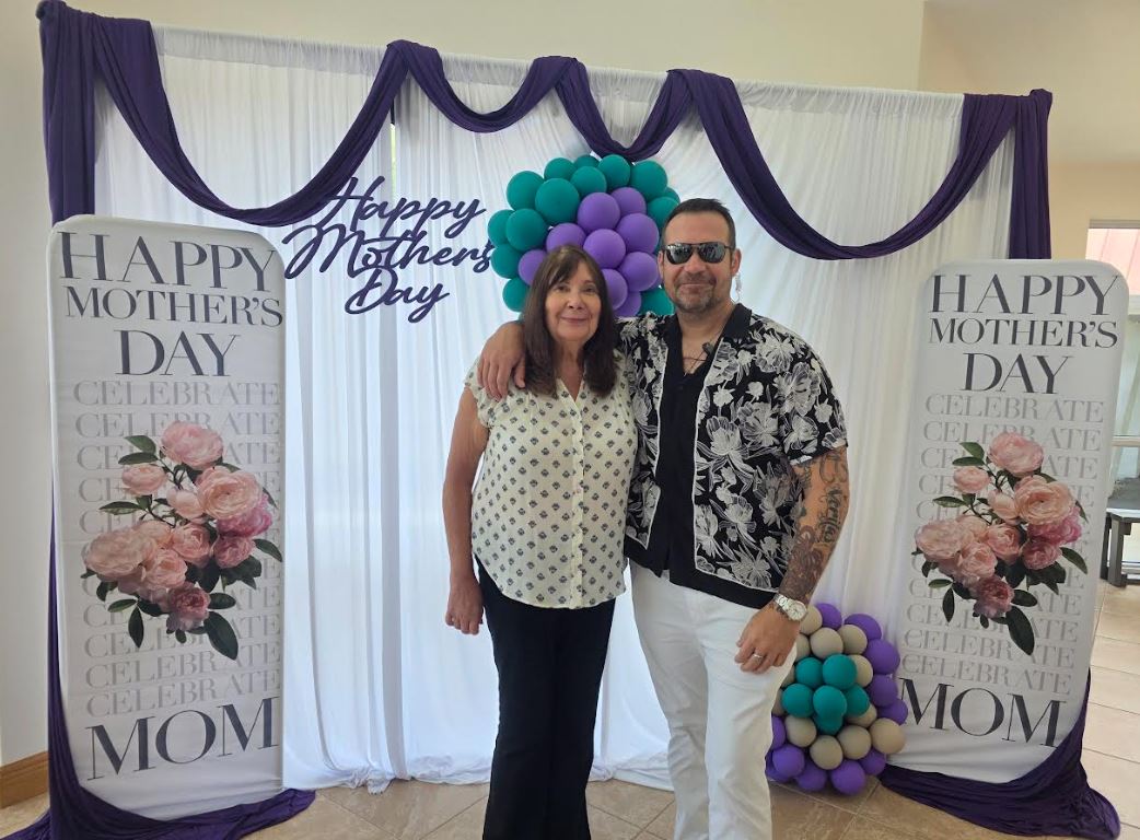 A man and woman pose together in front of a festive Mother's Day backdrop featuring floral decorations and colorful balloons.