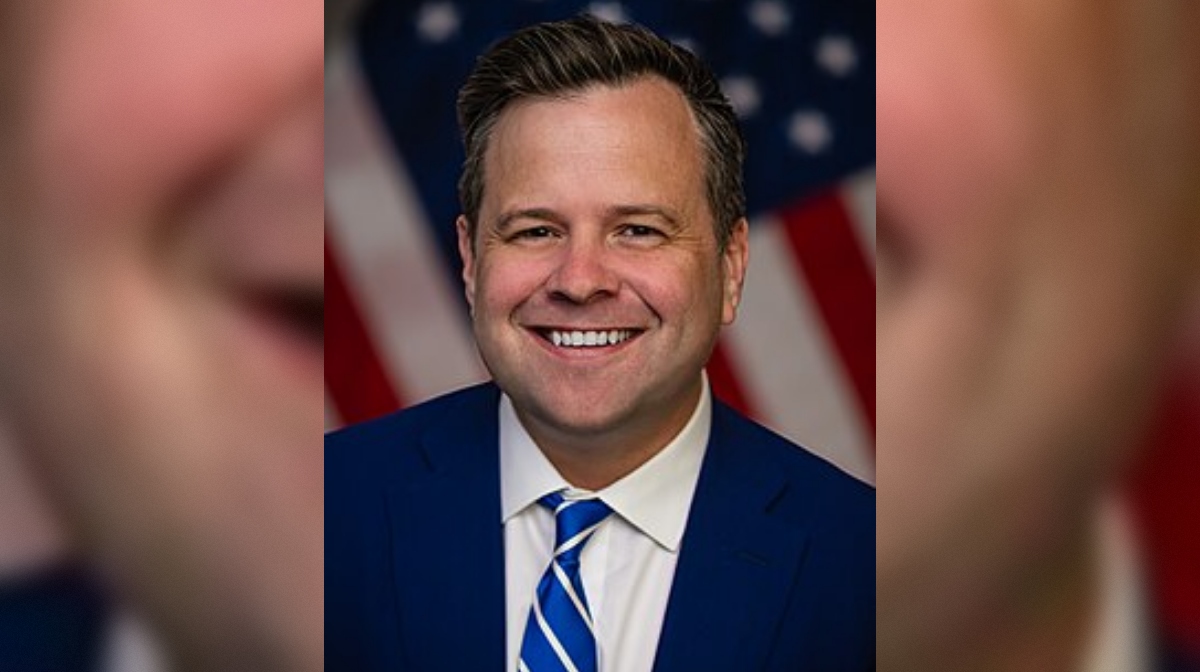 Professional headshot of a smiling man in a blue suit with a striped tie, set against a background of American flags.
