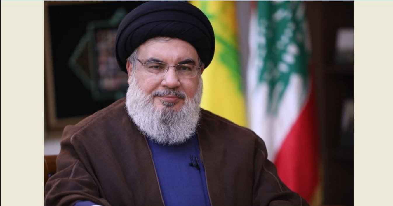Portrait of a man with a beard and glasses, wearing traditional attire, seated in front of Lebanese and Hezbollah flags.